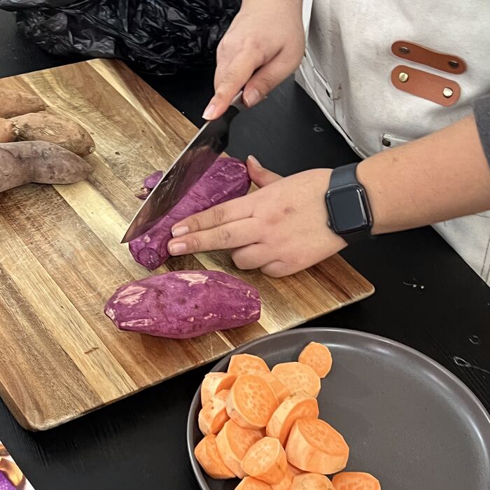 Hands slicing a purple ube on a wooden board next to a tray of sweet potato rounds